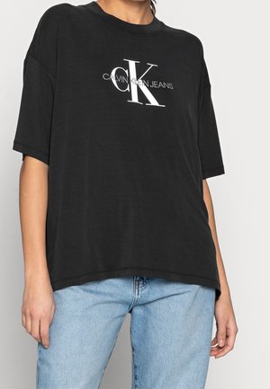 Personne portant un t-shirt noir ample avec le logo blanc "CK Calvin Klein Jeans" et un jean bleu clair sur un fond clair uni.