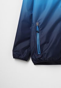 Marineblauwe windjack met een ombre-design, voorzien van een blauwe ritszak en elastische manchetten voor een stevige pasvorm. Soepel, lichtgewicht materiaal.