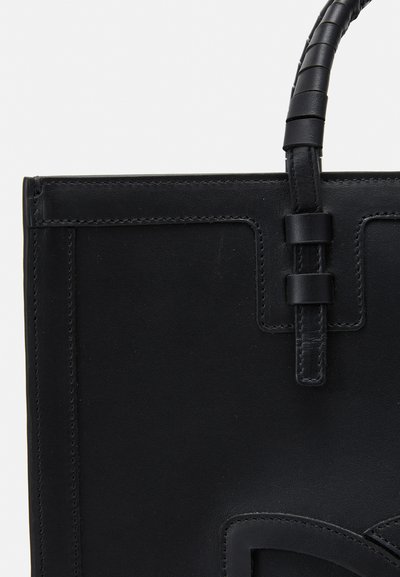 Sac à main en cuir noir présentant un design structuré, une poignée texturée et des coutures détaillées. Les accents notables comprennent un logo prominent et une finition lisse.
