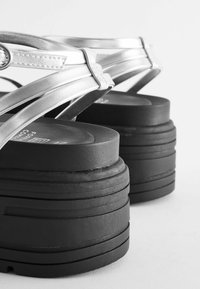 Juodi platform sandalai su storais, giliai galima riedėti padais ir metaliniais sidabriniais diržais, turintys tarpusavyje susijusį dizainą ir lygų paviršių.
