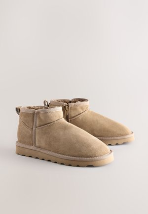 Beige ruskindsankelstøvler med lynlåslukning, blødt indvendigt foer og en tekstureret gummisål for godt greb. Har minimal syningsdetalje.