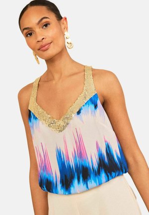 Sieviete valkā bezroku topu ar V-veida izgriezumu un zelta fliteru maliņu, zils, rozā un melns batikas raksts, kopā ar gaišas krāsas biksēm un zelta auskariem.