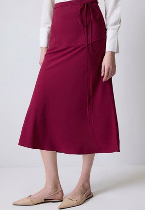 Jupe longue bordeaux en tissu doux, avec une taille haute, un détail de noeud à la ceinture et un ourlet évasé. Assortie à des chaussures légères à enfiler de couleur beige.