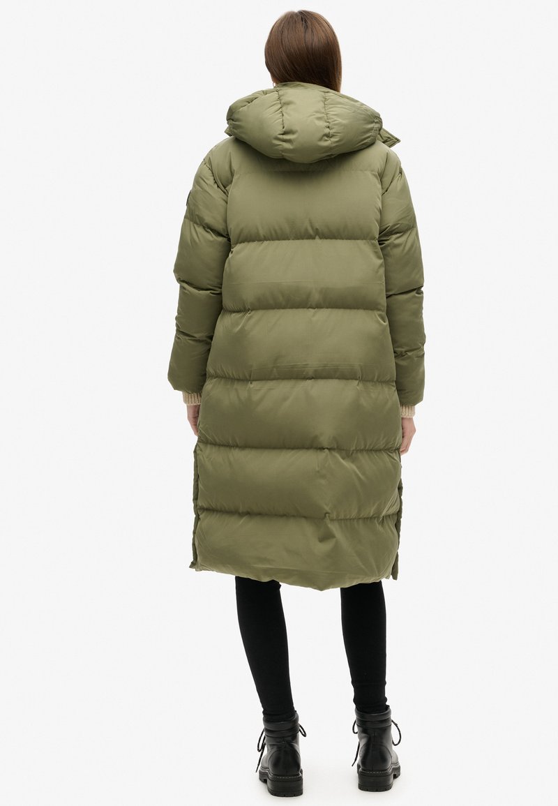 Olivgrüne Pufferjacke mit Kapuze, ausgestattet mit horizontaler Steppung, seitlichen Reißverschluss-Schlitzen und gerippten Bündchen. Sie reicht bis zur Mitte der Waden.