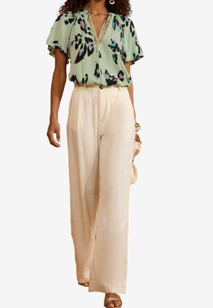 Femme portant un pantalon crème à jambes larges et un chemisier court vert à fleurs avec un col décoré, tenant un sac à main beige.