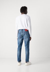 Celana jins denim biru dengan potongan lurus, menampilkan label HUGO merah di bagian belakang. Dipadukan dengan kemeja lengan panjang putih polos dan sneakers putih.