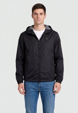 Młody mężczyzna ubrany w czarną kurtkę z kapturem na zamek błyskawiczny, na białej koszulce i niebieskich dżinsach, stojący na jasnym, jednolitym tle.