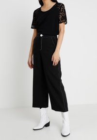 Svart blus med spetsärmar, tillsammans med högmidjade, vida svarta byxor med vit sömnad och vita ankelstövlar.