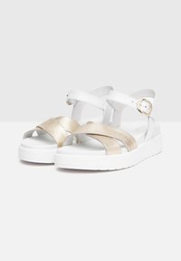 Sandales blanches et dorées à lanières avec une semelle blanche épaisse. Présente des bandes métalliques dorées sur le dessus du pied, une bride de cheville ajustable et une texture lisse.