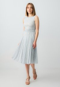 Helles blaues ärmelloses Kleid mit tailliertem Oberteil und ausgestelltem Rock, das mit einem karierten Muster in Gelb und Marineblau versehen ist. Getragen mit hellbraunen Sandalen.