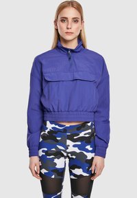 Lilla, kort jakke med høy krage, frontlomme og elastisk kant, sammen med camo leggings i flere farger med mesh-detaljer.