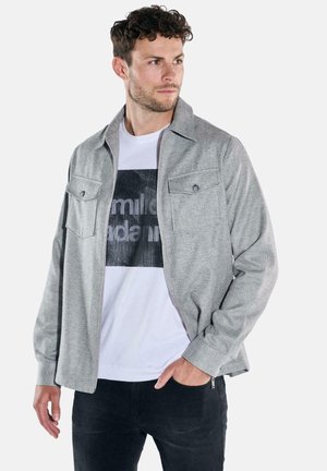 Grau strukturierte Jacke mit Kragen, zwei Brusttaschen und einem vorderen Reißverschluss. Getragen über einem weißen T-Shirt mit Grafik.