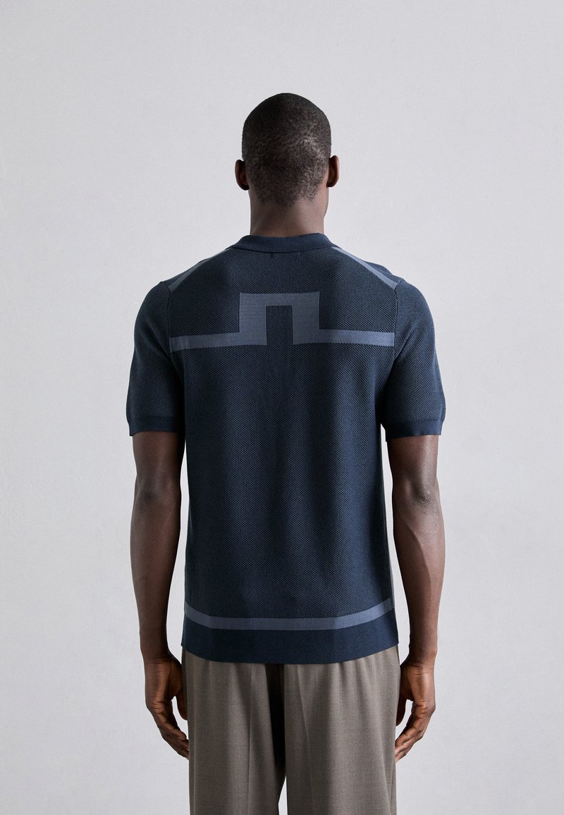Camiseta de punto de manga corta en azul marino oscuro con tejido texturizado. Presenta un diseño geométrico en azul más claro a lo largo de la espalda y un corte recto.