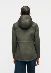 Personne avec des cheveux tressés portant une veste à capuche vert foncé et un jean bleu, vue de dos sur un fond uni.