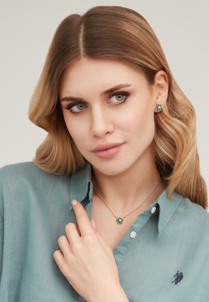 Donna con capelli biondi ondulati che indossa orecchini floreali verdi e una collana con ciondolo abbinata, vestita con una camicia verde petrolio con bottoni.