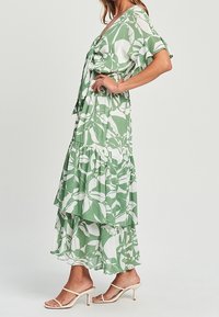 Vestido largo floral en verde y blanco, con mangas cortas, cintura ajustada con un lazo y volantes en las capas de la falda para una silueta fluida.