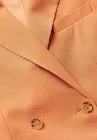 Dwurzędowa marynarka w kolorze brzoskwiniowym, wykonana z tkaniny o teksturowanym wykończeniu, z dwoma guzikami i kieszenią po lewej stronie. Prosty i nowoczesny design.