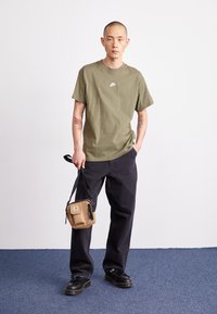 Olivza krāsas kokvilnas T-krekls ar mazu baltu Nike logo, apvienots ar melniem brīviem džinsiem un brūnu plecu somu. Melnas kedas.