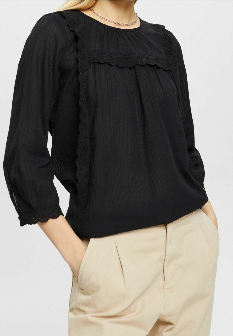 Schwarze Bluse mit strukturiertem Finish, gewellter Spitzenbesatz am Ausschnitt und dreiviertellangen Ärmeln. Kombiniert mit beigen Hosen.