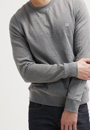 Mężczyzna ubrany w szarą, gładką bluzę z długim rękawem i małym logo na piersi, poprawiający mankiet, w zestawieniu z ciemnoniebieskimi dżinsami.