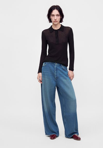 Femme aux cheveux courts et foncés portant un haut transparent noir à manches longues, un jean bleu à jambes larges et des chaussures bordeaux sur un fond uni.