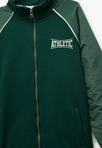 Dunkelgrüne Sportjacke mit Reißverschluss vorne, weißem Piping entlang der Nähte und "ATHLETIC" auf der Brust gestickt. Hat eine weiche Textur.