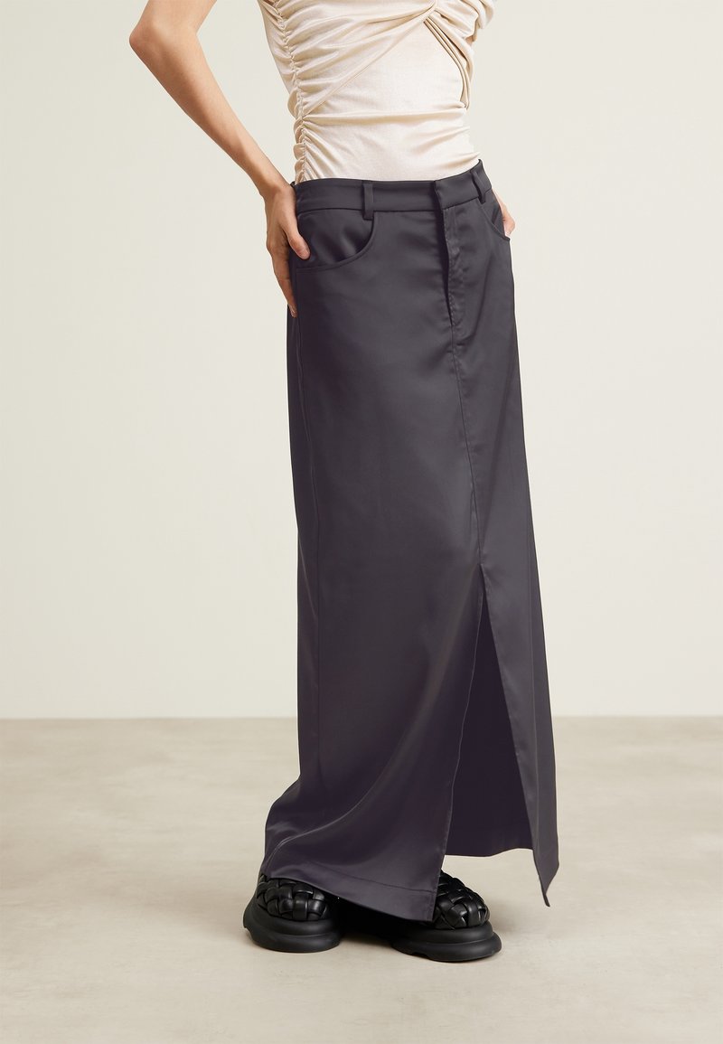 Kvinna i en mörkgrå lång kjol med slits fram, rynkad ljusbeige topp och svarta vävda platåskor som står på ett neutralt golv.