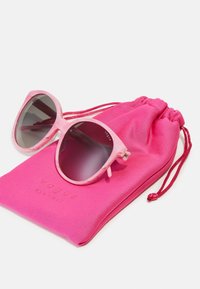 Ροζ διάφανα γυαλιά ηλίου με gradient σκούρους φακούς, τοποθετημένα πάνω σε ένα έντονο ροζ μαλακό θήκη με την επωνυμία "vogue eyewear" τυπωμένη με πεζά γράμματα.