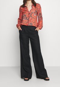 Blouse à imprimé floral rouge avec des manches longues et une fermeture à boutons, associée à un pantalon noir ample et des sandales plates. Matériau lisse et léger.