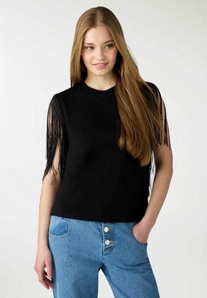 Giovane donna con capelli biondi lunghi che indossa una maglietta nera con frange a maniche corte e jeans azzurri a vita alta, con la mano in tasca, sorridente.