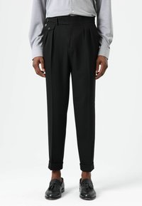 Zwarte broek met plooien, voorzien van een met knopen gesloten zijzak en omgeslagen zoom, gecombineerd met een lichtgrijs shirt met lange mouwen en zwarte schoenen.