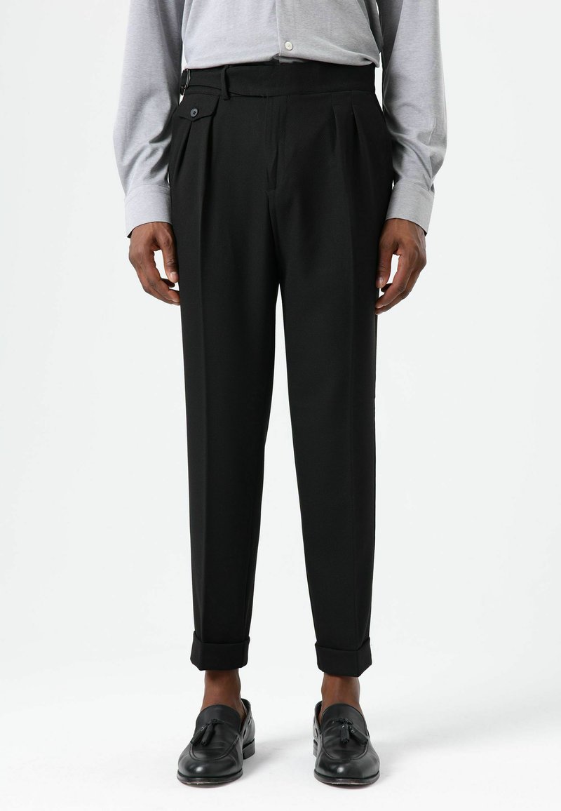 Zwarte broek met plooien, voorzien van een met knopen gesloten zijzak en omgeslagen zoom, gecombineerd met een lichtgrijs shirt met lange mouwen en zwarte schoenen.