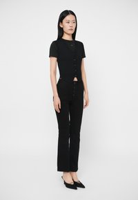 Γυναίκα που στέκεται φορώντας μαύρο κοντομάνικο τοπ, μαύρη γουνάκι με κουμπιά, μαύρα ψηλόμεσα τζιν και μαύρες μυτερές γόβες σε λευκό φόντο.