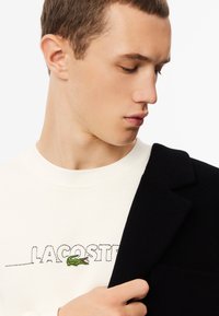 Felpa bianca con logo "LACOSTE" ricamato in nero e coccodrillo verde, abbinata a un cappotto di lana nero. Texture liscia, design su misura.