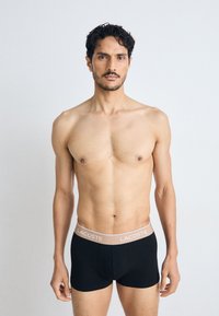 Boxers noirs avec une taille de couleur marron clair embossée du logo "LACOSTE." Le tissu semble lisse et extensible, épousant étroitement le corps.
