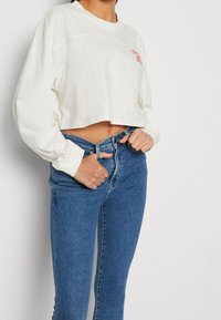 Sudadera blanca de manga larga, cortada, con un estampado en el pecho, combinada con jeans ajustados azules. Tejido suave, corte relajado y diseño sencillo.