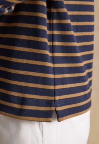 Ourlet de chemise rayé bleu marine et moutarde avec fente latérale, porté sur un pantalon blanc, montrant la texture du tissu et les détails des coutures.
