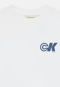 Felpa bianca in cotone con scollatura rotonda, con il logo blu "CK" sul petto a sinistra e un'etichetta con scritto "Calvin Klein Jeans."