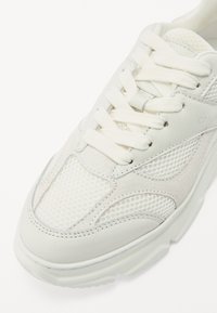 Zapatilla blanca con una parte superior de malla texturizada, acentos de cuero suave y cordones planos. Presenta una suela de goma gruesa y detalles de costura sutiles.