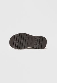 Suela de zapato de goma negra con patrones de huella profundos y surcos para tracción; punta redondeada suave y un logotipo de marca prominente en el centro.