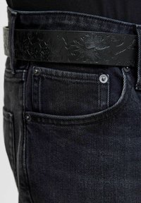 Acercamiento a un bolsillo de jeans de mezclilla oscura con remaches metálicos y un cinturón de cuero negro con un grabado floral.