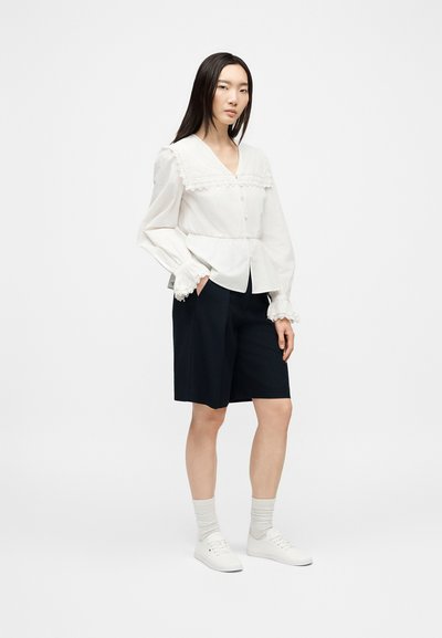 Femme portant une blouse blanche à manches longues avec des détails en dentelle, un short noir au-dessus du genou, des chaussettes blanches et des baskets blanches, debout les mains dans les poches.