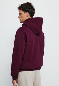 Felpa con cappuccio borgogna, dal tessuto morbido, vestibilità rilassata e polsini a costine, mostrata da dietro senza loghi o motivi visibili.