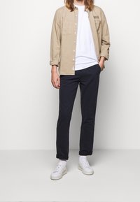 Beige overhemd met knopen over een wit T-shirt, gecombineerd met marineblauwe broek en witte sneakers. Het overhemd heeft twee borstzakken en opgerolde mouwen.