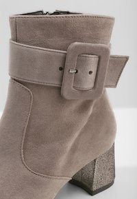 Botín de ante en beige claro con una correa ancha y hebilla rectangular. Cuenta con un tacón cuadrado texturizado para un detalle adicional.