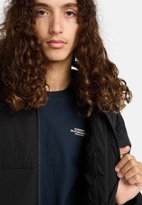 Sort jakke med lynlås og sidelommer, båret over en navyblå t-shirt med hvid tekst. Modellen har krøllet brunt hår.