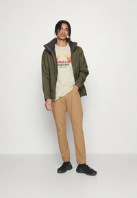 Zöld vízálló dzseki szürke béléssel, bézs színű, hegy mintás grafikus póló, világosbarna nadrág és fekete sportcipő.