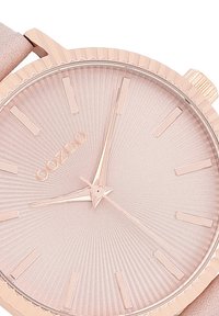 Montre ronde en or rose avec un cadran rose pâle texturé, des index horaires fins en or rose et un bracelet en cuir rose lisse. Nom de la marque "OOZOO" affiché.