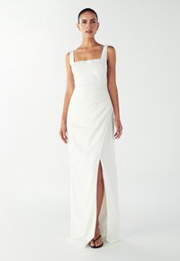 Žena nosi bezrukavnu bijelu haljinu do poda s kvadratnim dekolteom i prorezom na butini, u kombinaciji s crnim sandalama.