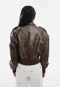 Personne aux longs cheveux tressés portant une veste aviateur en cuir marron et un pantalon blanc, vue de dos sur un fond uni.